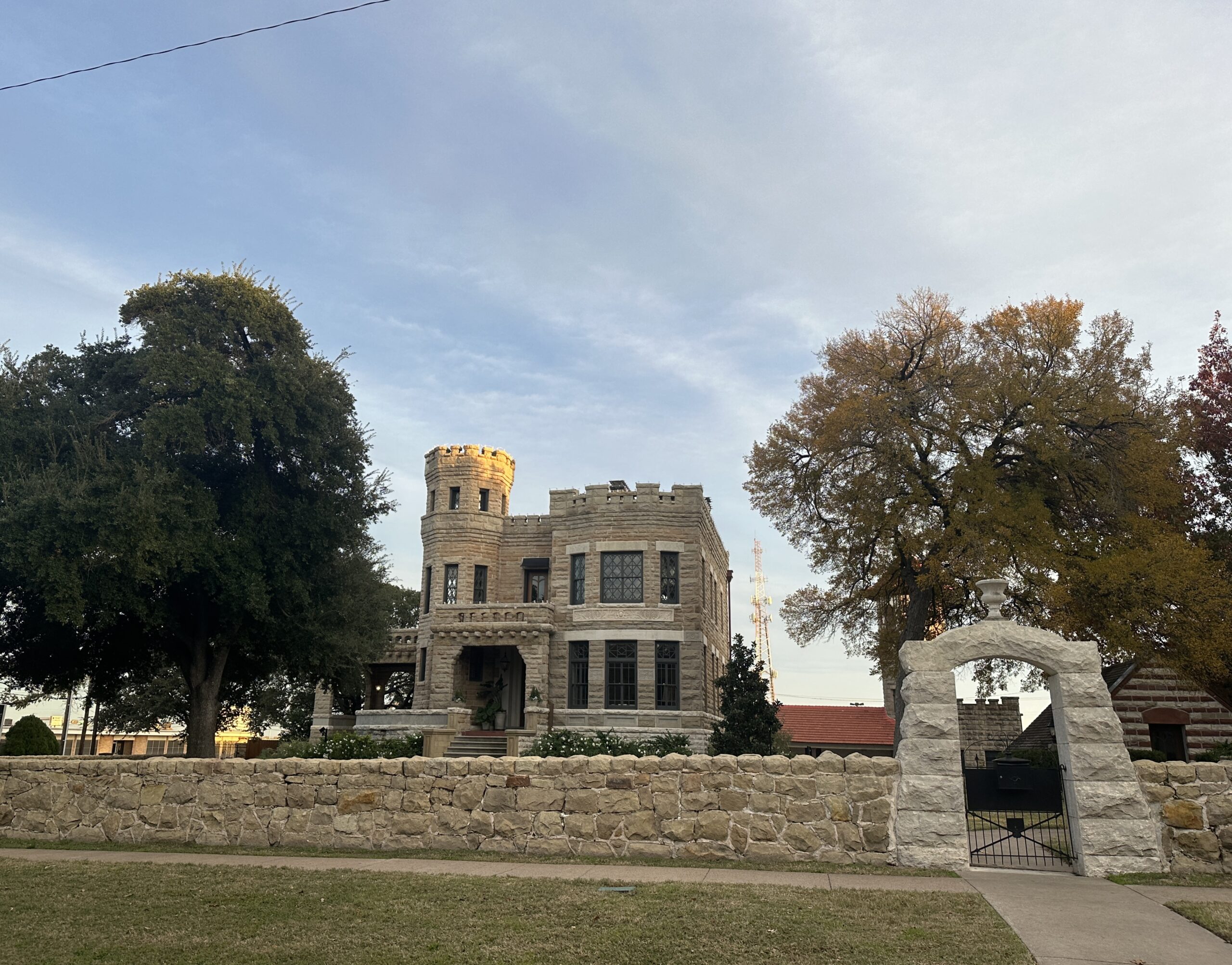 A castle that is made of white stone with a gate surrounding the complex. It's in Waco, Texas. The sky is blue with few clouds. The grass is tinted yellow, but the trees are green.